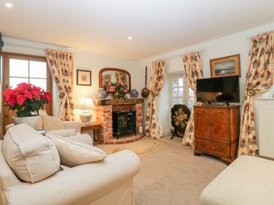 A living room with a beige sofa poinsettia plant a brick fireplace with a mirror floral curtains and a wooden cabinet with a television at Colly Cottage in Dottery