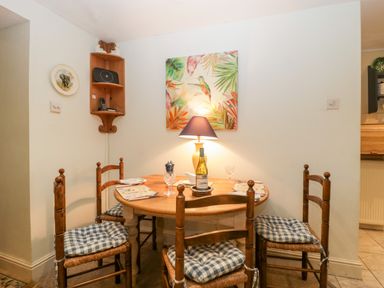 A dining area with a round wooden table and four wooden chairs with checkered cushions a table lamp and wall art above at Colly Cottage in Dottery