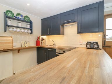 A kitchen with wooden countertops dark blue cabinets a sink a stovetop toaster and a shelf with cups and containers at Colly Cottage in Dottery