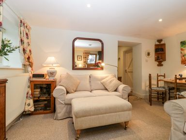 A living room with a beige sofa and ottoman a wooden side table with a lamp and books and a wooden dining table with chairs at Colly Cottage in Dottery
