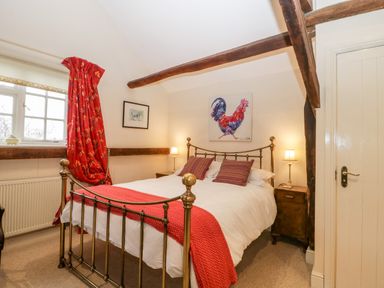 A bedroom with a metal bed frame red throw pillows and a red blanket beside a window with red curtains and wooden beams at Colly Cottage in Dottery