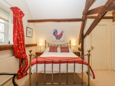 A bedroom with a brass bed frame red blanket and curtains wooden beams and a rooster painting above the bed at Colly Cottage in Dottery