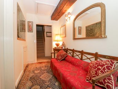 A hallway with a red patterned sofa a wooden side table with a lamp wall mirror and staircase at Colly Cottage in Dottery