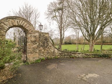 A stone wall with an arched iron gate and a lamp post next to a paved area and trees in a field at Colly Cottage in Dottery