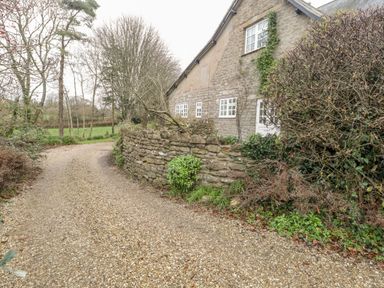 A gravel driveway curving around a stone wall with shrubs and a stone house with white windows in the background at Colly Cottage in Dottery