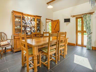 A dining room with a wooden table and six chairs a wooden cabinet with dishes and mugs a wooden door and window with floral curtains at Ysgubor Hir Pontarddulais near Swansea