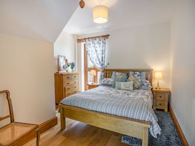 A bedroom with a wooden bed a striped and floral bedspread pillows a wooden dresser with a mirror and flowers a wooden chair and a window with a floral curtain at Ysgubor Hir in Pontarddulais near Swansea