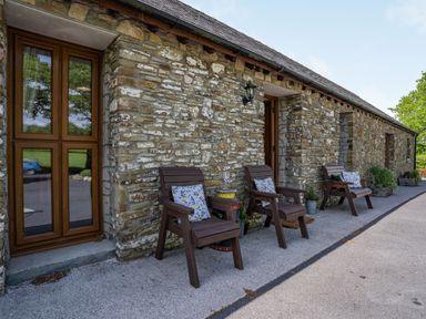 An outdoor seating area with three wooden chairs with cushions and a stone building at Ysgubor Hir in Pontarddulais near Swansea
