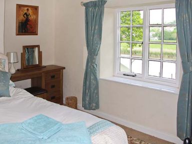 A bedroom with a bed and a wooden dressing table next to a window with blue curtains at Tuckermarsh Quay River Cottage 2 Bere Alston near Tavistock