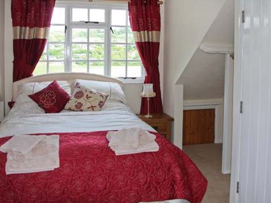 A bedroom with a bed covered with red and white bedding and two pillows next to a window with red curtains at Tuckermarsh Quay River Cottage 2 Bere Alston near Tavistock