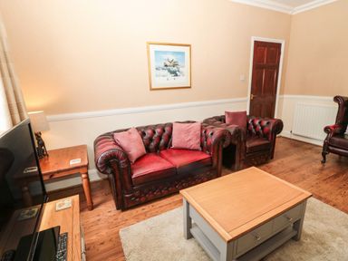 A living room with red leather sofas wooden coffee and side tables and a television at Coquet Cottage in Amble