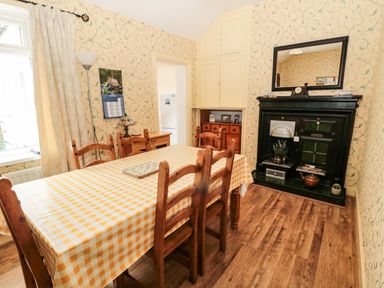 A dining room with a table covered by a yellow checkered tablecloth wooden chairs a black fireplace with a mirror above and a wooden floor at Coquet Cottage in Amble
