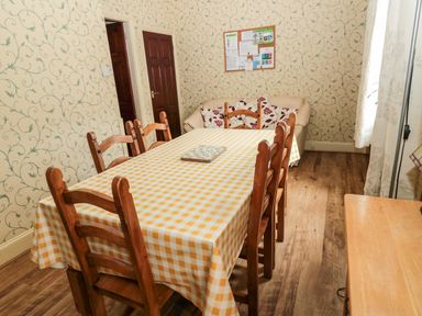A dining room with a wooden table covered with a yellow checkered tablecloth and six wooden chairs a cream sofa with patterned cushions a corkboard on the wall wooden floor and patterned wallpaper at Coquet Cottage in Amble
