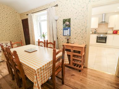 A dining room with a wooden table and six chairs next to a window and a small cabinet next to a doorway leading to a kitchen at Coquet Cottage in Amble