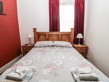 A bedroom with a wooden bed frame floral bedding two bedside tables with lamps and red curtains at Coquet Cottage in Amble