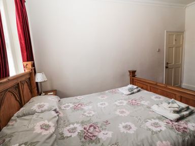 A bedroom with a wooden bed, floral bedding, red curtains, a lamp on a small bedside table, and towels on the bed at Coquet Cottage in Amble