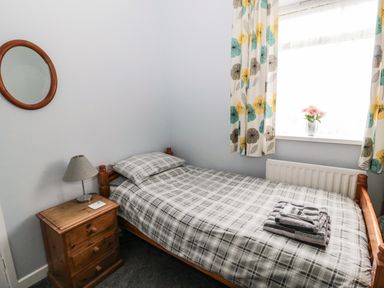 A bedroom with a single wooden bed with checked bedding a wooden nightstand with a lamp a round wall mirror and a window with curtains and a vase of flowers at Coquet Cottage in Amble