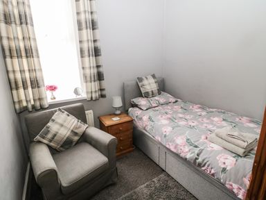 A small bedroom with a single bed with floral bedding a gray armchair a wooden nightstand with a lamp pink flowers on the windowsill and plaid curtains at Coquet Cottage in Amble