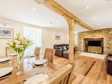 A dining area with a wooden table and chairs set for six people next to a living room with leather sofas and a stone fireplace at Ysgubor Uchaf Brongest near Newcastle Emlyn