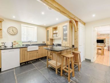 A kitchen with wooden cabinets stone floor a clock on the wall and two wooden stools at Ysgubor Uchaf in Brongest near Newcastle Emlyn