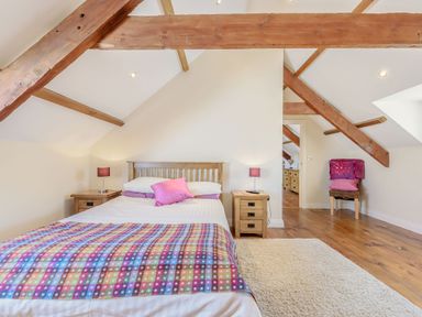 A bedroom with a wooden bed and colorful blanket and pillows two bedside tables with lamps and a chair with cushions under exposed wooden beams at Ysgubor Uchaf Brongest near Newcastle Emlyn