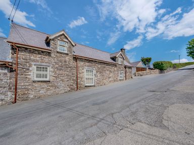 A stone house with white windows on a sloped paved road under a blue sky at Ysgubor Uchaf in Brongest near Newcastle Emlyn