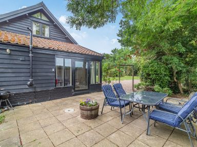 An outdoor patio with a glass table and four chairs with blue cushions outside a black wooden house at Ducksfoot Barn Bush Green near Pulham Market