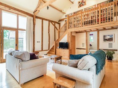 A living room with two beige sofas a wooden coffee table a TV on a wooden cabinet stairs leading to a bookshelf balcony and large windows at Ducksfoot Barn in Bush Green near Pulham Market