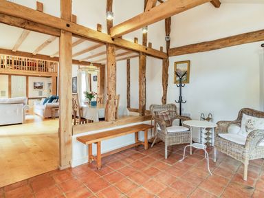An open living and dining area with wooden beams two wicker chairs a small white table and a wooden bench at Ducksfoot Barn in Bush Green near Pulham Market