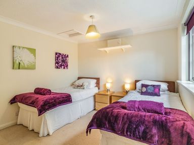 A bedroom with two single beds with purple blankets and pillows two bedside tables with lamps and floral wall art at Ducksfoot Barn in Bush Green near Pulham Market