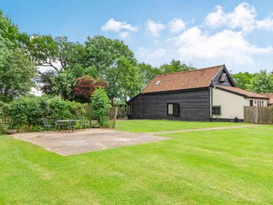 A black wooden house with a tiled roof and a patio set with chairs on a paved area surrounded by grass at Ducksfoot Barn in Bush Green near Pulham Market