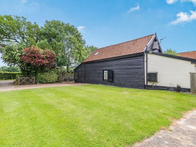 A garden with green grass and trees beside a black wooden barn with a brown tiled roof at Ducksfoot Barn in Bush Green near Pulham Market