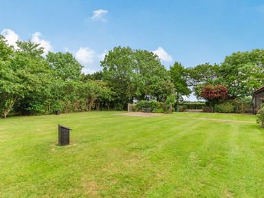 A large grass lawn with trees and bushes around and a small paved area with chairs at Ducksfoot Barn in Bush Green near Pulham Market