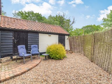 An outdoor patio area with two chairs and a small table near a brick and pebble ground beside a building with a tiled roof and wooden doors at Ducksfoot Barn in Bush Green near Pulham Market