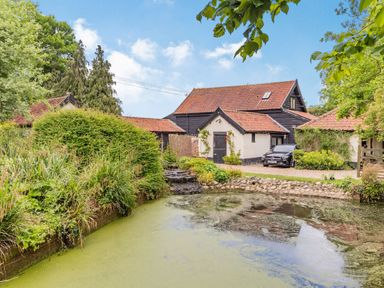 A pond with plants and a house with tiled roof and car in the driveway at Ducksfoot Barn Bush Green near Pulham Market