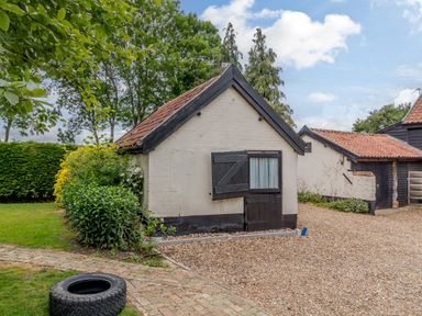 A small white brick building with a tiled roof and black door surrounded by gravel and greenery at Garden Cottage in Bush Green near Pulham Market