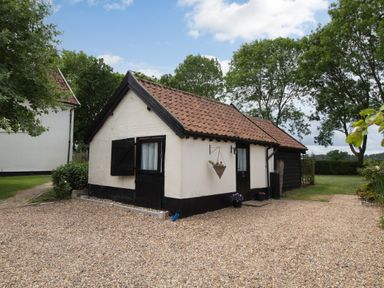 A small white cottage with black trim and a tiled roof surrounded by trees and lawn at Garden Cottage in Bush Green near Pulham Market