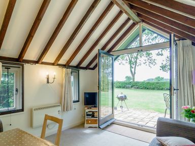 A room with wooden beams a dining table chairs a TV stand a sofa and an open glass door leading to a garden at Garden Cottage Bush Green near Pulham Market