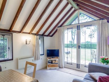 A living room with exposed wooden beams a dining table a television on a stand and glass doors overlooking a garden at Garden Cottage Bush Green near Pulham Market