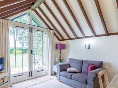A living room with a gray sofa a side table with flowers a floor lamp and glass doors leading to a garden at Garden Cottage in Bush Green near Pulham Market