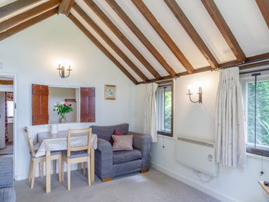 A living room with wooden beams on the ceiling a small dining table with chairs an armchair two windows with curtains and wall lamps at Garden Cottage in Bush Green near Pulham Market