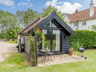 A small black wooden cottage with a tiled roof and glass doors in a garden with patio furniture and a barbecue at Garden Cottage in Bush Green near Pulham Market