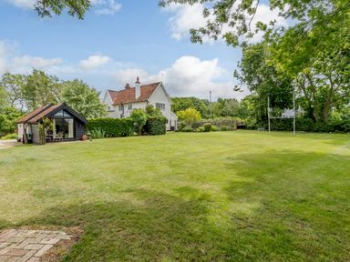 A large grass lawn with a small black garden shed and a white house with a red tiled roof at Garden Cottage in Bush Green near Pulham Market