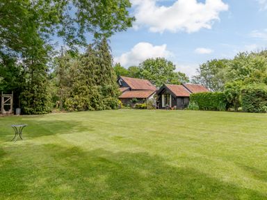 A large grassy lawn with trees and two black wooden buildings with red roofs at Garden Cottage in Bush Green near Pulham Market
