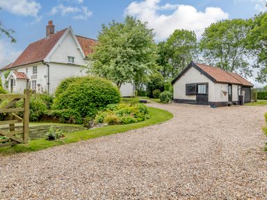 A gravel driveway leading to a white house and a smaller outbuilding with trees and shrubs at Garden Cottage in Bush Green near Pulham Market