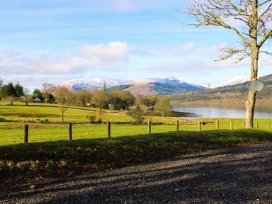 A landscape view of a grassy field with a gravel path a tree and a lake with snow capped mountains in the background at The Four Oaks Chalet in Blaich near Fort William