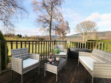 A wooden deck with outdoor wicker furniture and a bottle of wine overlooking a grassy landscape at The Four Oaks Chalet in Blaich near Fort William