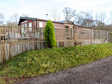 Wooden chalet with a deck and outdoor furniture surrounded by grass and trees at The Four Oaks Chalet in Blaich near Fort William