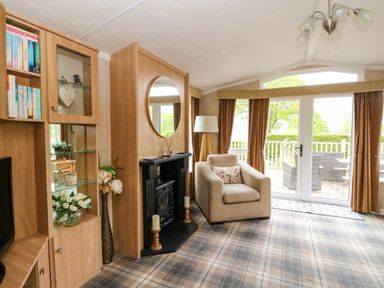 A living room with a beige armchair and fireplace next to glass doors and windows at The Four Oaks Chalet in Blaich near Fort William