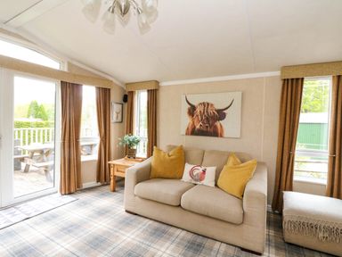 A living room with a beige sofa and yellow cushions a wooden side table and windows with brown curtains at The Four Oaks Chalet in Blaich near Fort William